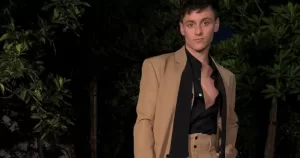 The image shows sports star Tom Daley, posed against a background of shrubbery, wearing a brown suit with an open black shirt. His latest YouTube vlog posted earlier this week.