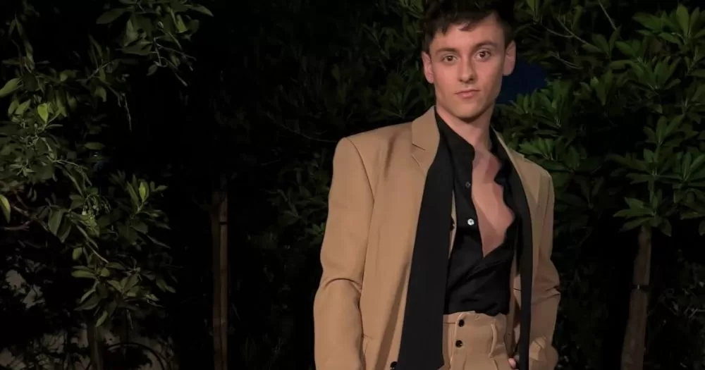 The image shows sports star Tom Daley, posed against a background of shrubbery, wearing a brown suit with an open black shirt. His latest YouTube vlog posted earlier this week.