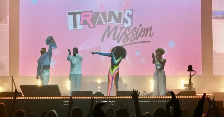 Four performers stand on a stage. The pink backdrop reads 'Trans Mission'.