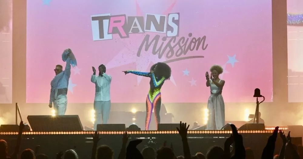 Four performers stand on a stage. The pink backdrop reads 'Trans Mission'.