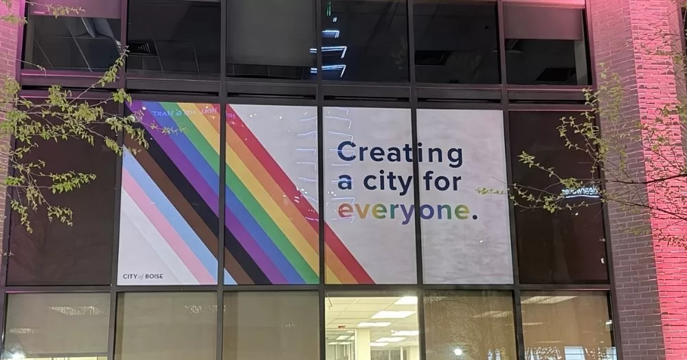A banner can be seen through a window at Boise City Hall. The poster reads "Creating a city for everyone," and there is a stripe of colours from the Progress Pride flag running across the banner.