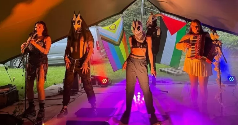 Artists performing at Bród na Gaeltachta, inside a tent. They are wearing masks and have a Pride flag and a Palestinian flag in the background.