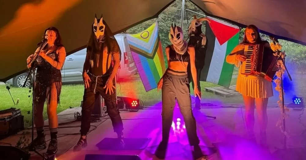 Artists performing at Bród na Gaeltachta, inside a tent. They are wearing masks and have a Pride flag and a Palestinian flag in the background.