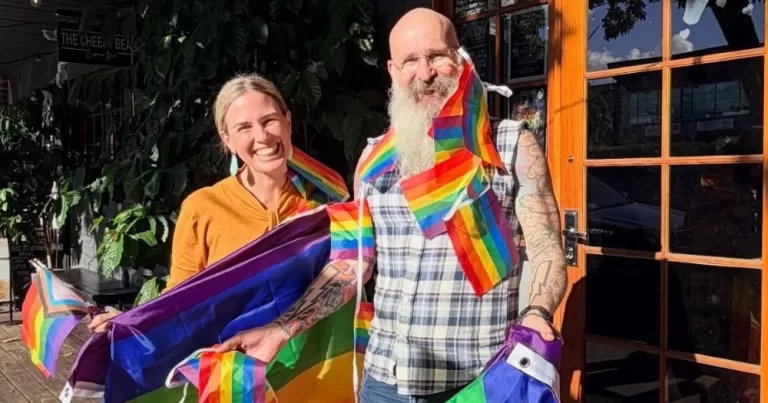 The picture shows Billerwell Daye, the owner of Come to Daddy bar in Brisbane, wrapped in pride flags and standing next to MP Renee Coffey as she comes to support the bar after an arson attack.