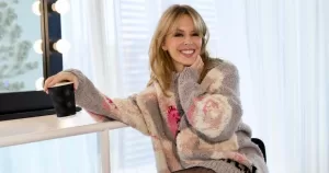 A photograph of Kylie Minogue next to a mirror, wearing a smile and a lovely cozy-looking jumper. Kylie Minogue documentary announced.