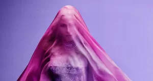 A cropped version of the cover art for Madonna's new album Confessions II. She poses in front of a purple background with a pink veil over her face.