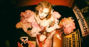 SexyTadhg announces new tour, SLUTTRAD. SexyTadhg poses with a violin and smoulders wearing a pink outfit nestled amongst pink feather boas and wigs.