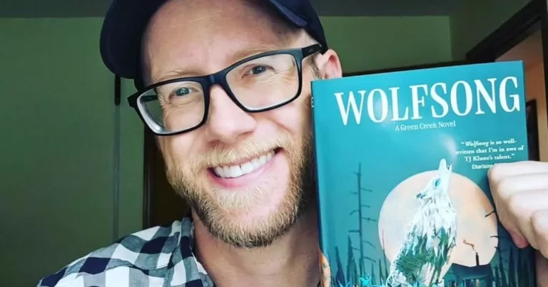 Author TJ Klune holding up his book 'Wolfsong' and smiling at the camera.