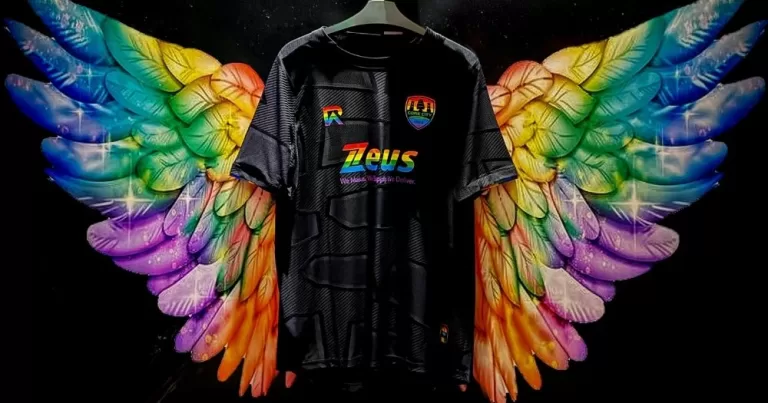 The image show's Cork City FC's newest LGBTQ+ inspired jersey, which is black with rainbow-coloured logos and club crest. The jersey is against a black wall background with rainbow-painted wings emerging from each side.
