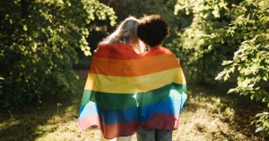Two people embrace wrapped in a Pride flag. This article covers a new resource aiming to support LGBTQ+ sobriety.