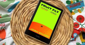 A kindle with the cover of novel Fruit Fly, by Josh Silver, with a bright green background, an orange and a fruit fly.