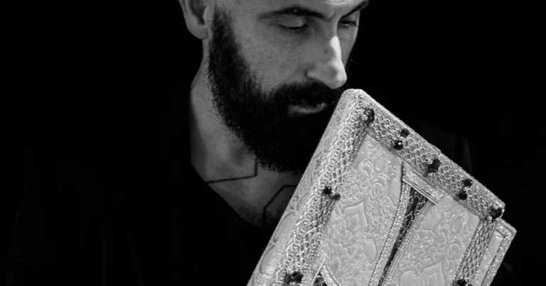 This black-and-white image shows gay designer Filipo Sorcinelli, who creates clothes for Pope Leo XIV. In the image he is looking down to the right, holding a manuscript in his hands, and wearing a black shirt.