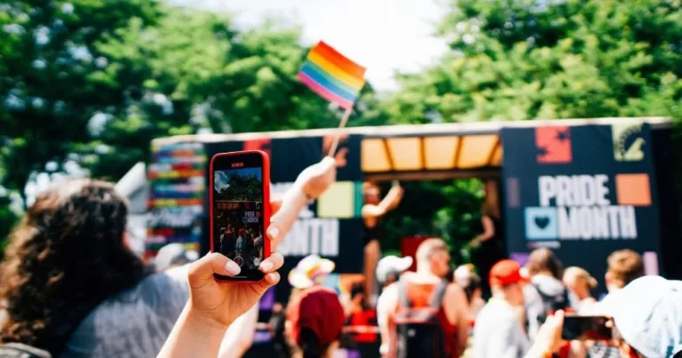The image shows a Pride event, to represent Hungarian voters' desire for greater LGBTQ+ rights. At the centre of the image is a crowd gathered in front of a open stage truck, with someone holding a Pride flag up high, and a person close to the camera recording with their phone.