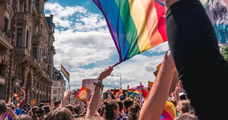 IDAHOBIT 2026. A large crowd is marching down a busy street, the point of view of the camera is in the middle of the crowd. In front of the camera is an arm upstertched waving a pride flag