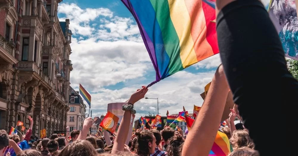 IDAHOBIT 2026. A large crowd is marching down a busy street, the point of view of the camera is in the middle of the crowd. In front of the camera is an arm upstertched waving a pride flag