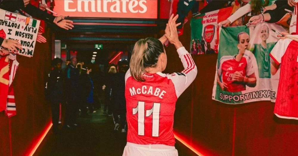The image shows Katie McCabe leaving Emirates stadium pitch. We see McCabe's back and her Arsenal jersey number 11. She is raising her hands to clap at the crowds, which are leaning over the barriers to cheer her on.