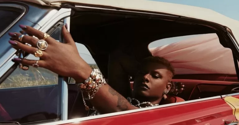 Liniker's album cover for Caju. Liniker sits reclined in a car's front seat, with her hand wearing several rings resting on the outside of the window.