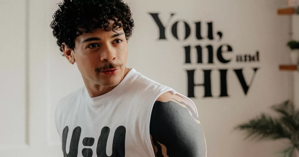 HIV activist Phellipe, who is from Brazil and hosted an episode of Poz Vibe podcast, posing for a photo while wearing a U=U t-shirt, with a sign in the background that reads "You, me and HIV".