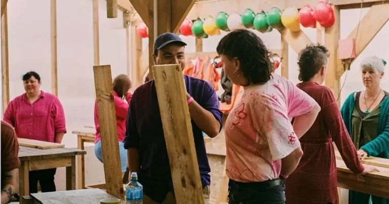 This article is about a Queer Sheds funded residency. Multiple people are standing talking to each other in a large tent, they stand in pairs in front of wooden tables. In the foreground are two people facing each other holding large wooden planks.