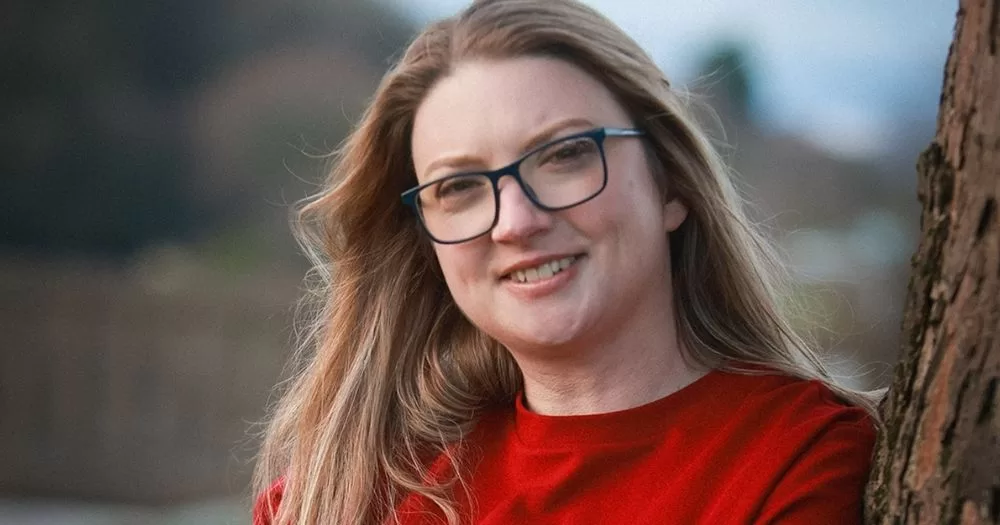 Portrait of Rebecca Murphy. She is standing outdoors with a red top and glasses on, and smiles at the camera.