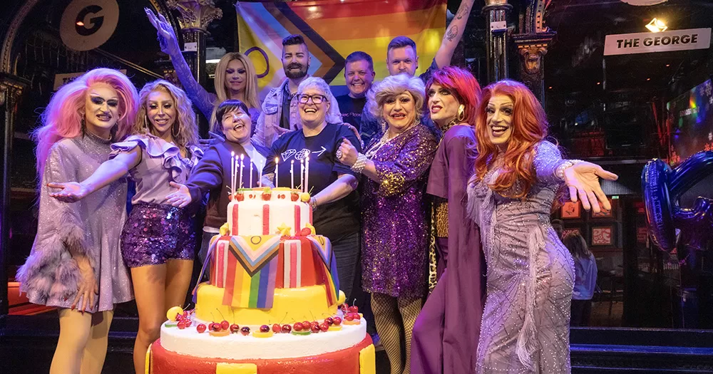 Drag artists, DJs and staff from The George pose on stage with a cake and rainbow flag.