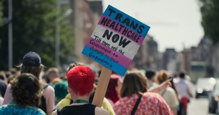 This article is about a new trans healthcare approach. In the photo, a person marching in a crowd in Dublin, carrying a sign that reads "Trans healthcare now".