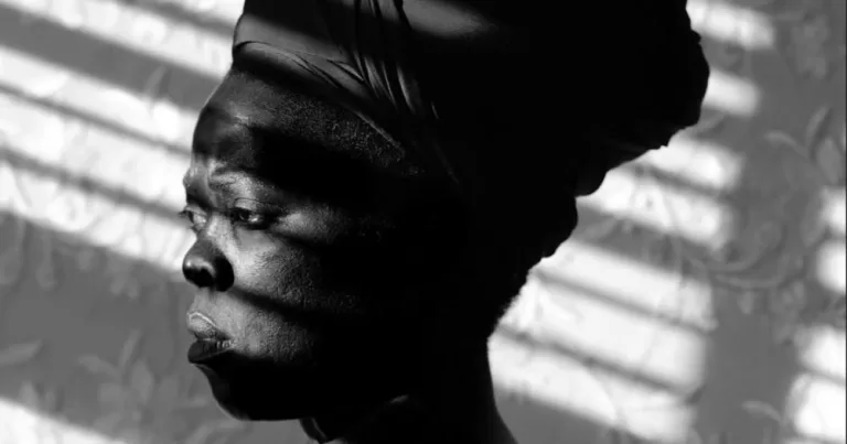 Black and white portrait of Non-binary photographer, Zanele Muholi. They are looking off to the side away from the camera, there is rays of light shining on their face, they are wearing a headdress