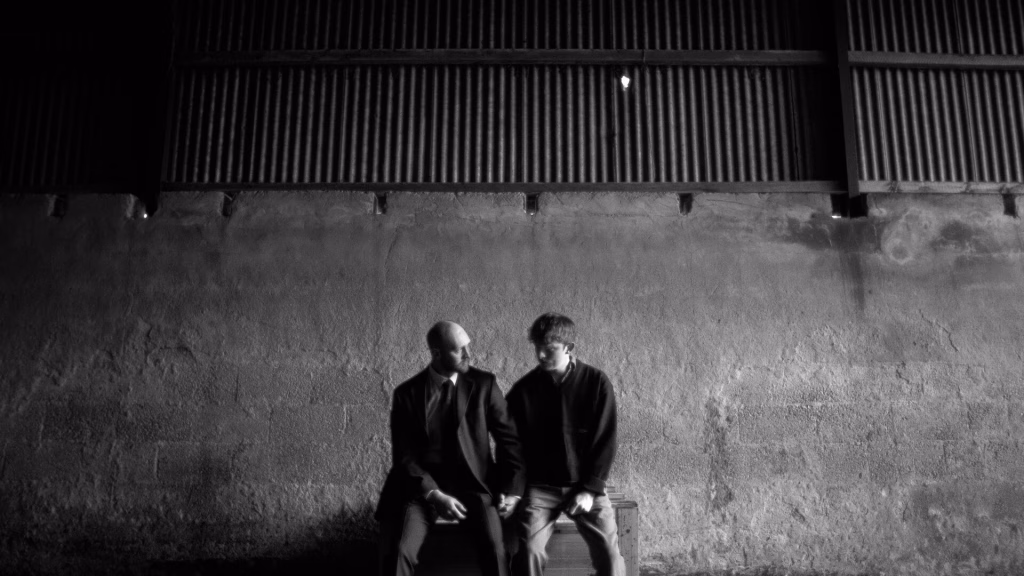 Black and white image of two men sitting on a bench looking at each other.