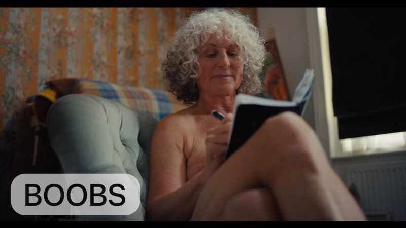 An older woman sitting naked on a chair reading a book.