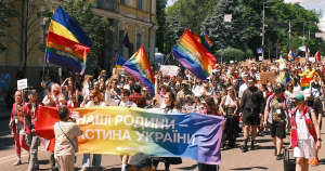 The crowd at the 10th Kyiv Pride march. People hold a rainbow banner and rainbow flags as they walk through the city.