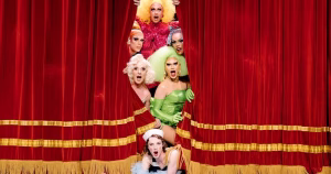This is an image of the Mockie Ah! drag collective on a stage. They are situated in the middle of a red stage curtain.