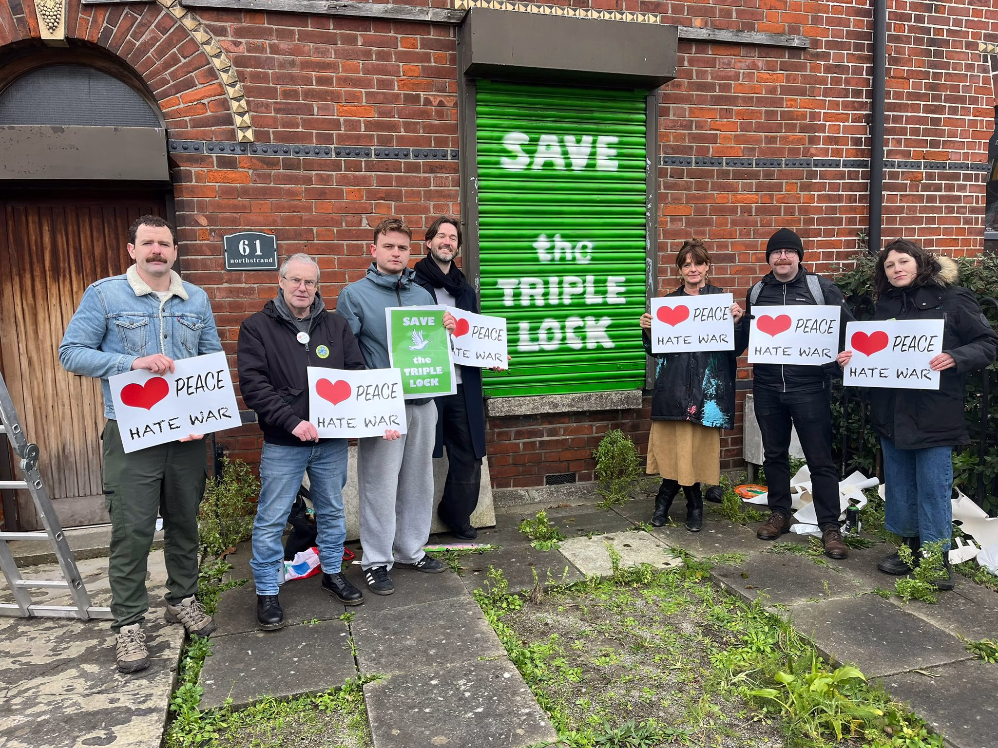 This article is about Irish neutrality. In the photo, activists standing in front of a wall with the message "Save the Triple Lock" holding banners that read "Love peace, hate war".