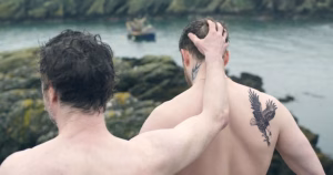 This is a still from queer film On The Sea. The image depicts the two main characters. They are shirtless, looking out at the water while one character puts his arm on the other's head.