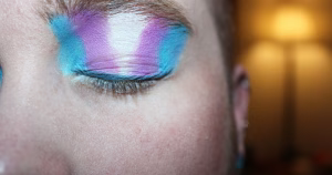 This article is about passing culture in the trans community. The image is a close-up of a person closing their eye, showing eyeshadow in the colours of the trans flag