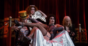 Still image from The Rocky Horror Picture Show of character Frank-N-Furter sitting on a sliver throne surrounded by other characters Riff Raff, Magenta, Columbia.