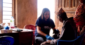 This article is about US funding cuts. Photo shows a volunteer working in a clay house, providing medical treatment to a man sitting on the floor.