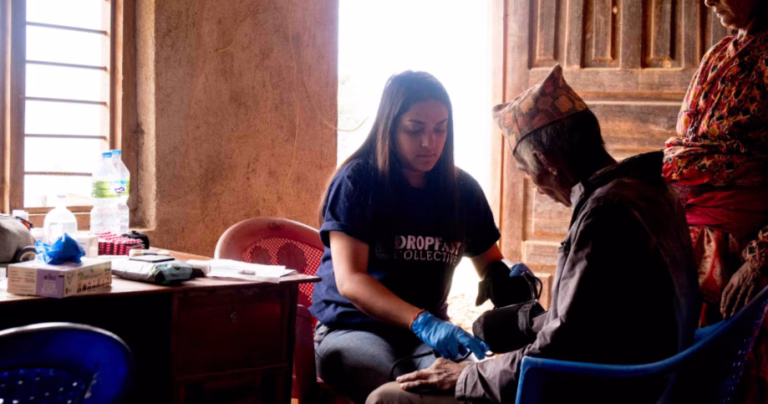 This article is about US funding cuts. Photo shows a volunteer working in a clay house, providing medical treatment to a man sitting on the floor.