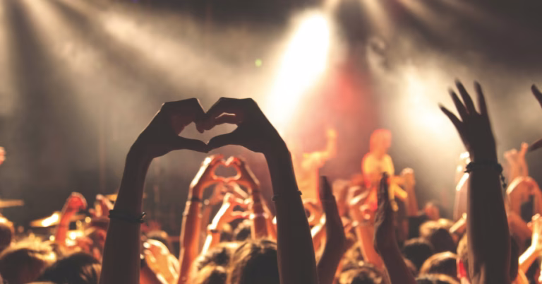 This article is about LGBTQ+ events happening in August. This image shows a crowd, illuminated with warm spot lights. The focal point of the image is someone making a heart with their hands in the air
