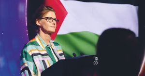 Anna Nolan at a podium on stage at The GALAS. The Palestinian flag is visible in the background.