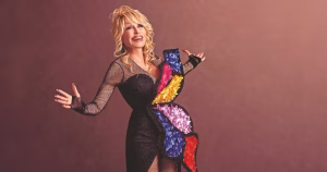 An image of Dolly Parton in front of a brown/red background. She smiles with her arms wide and looks into the distance.