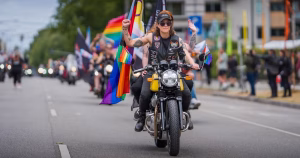 An image of two Dykes on Bikes members riding down a street with a Pride flag on the back of their bike.