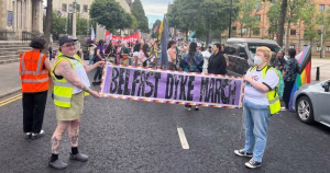 Two people in high vis vests holding a banner that reads 'Belfast Dyke March'. There is a crowd of marchers behind them in the background.