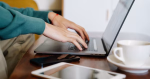 GCN is seeking an Online Content Creator. The image shows a close up of a person's hands working on a laptop. There is a mug and teacup beside it and an iPad and phone.
