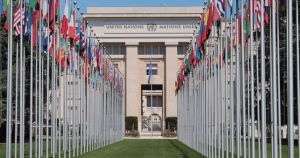 This article is about Irish civil society organisations highlighting Ireland's human rights shortcomings in Geneva. The image shows the exterior of the United Nations building with a row of flags in the foreground.