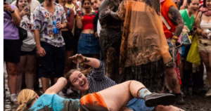 The article discusses the lesbian mud-wrestling competition at Lollapaloza. The image depicts two women wrestlng in mud with a crowd in the background