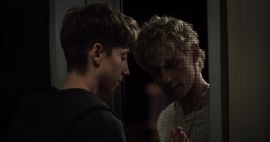 The image is a still from the film Leviticus. It shows two characters holding their hands together through a glass door.
