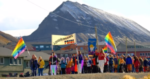 An image from the world's northernmost Pride in Longyearbyen. A crowd of people walk with flags and banners and an snow-capped mountain can be seen in the background.