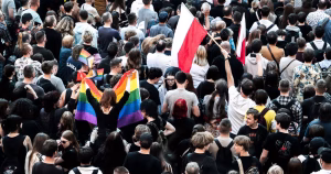 The image depicts a crowd rallying, people holding a Pride flag and Poland flag.