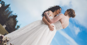 Two women in white wedding dresses holding each other. There is a blue sky in the background.