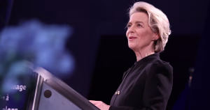 This article is about a proposed EU social media ban. European Commission President Ursula Von der Leyen speaking from a podium.